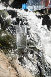 Photo of Woman pouring water from plastic bottle into glass near stream outdoors, closeup