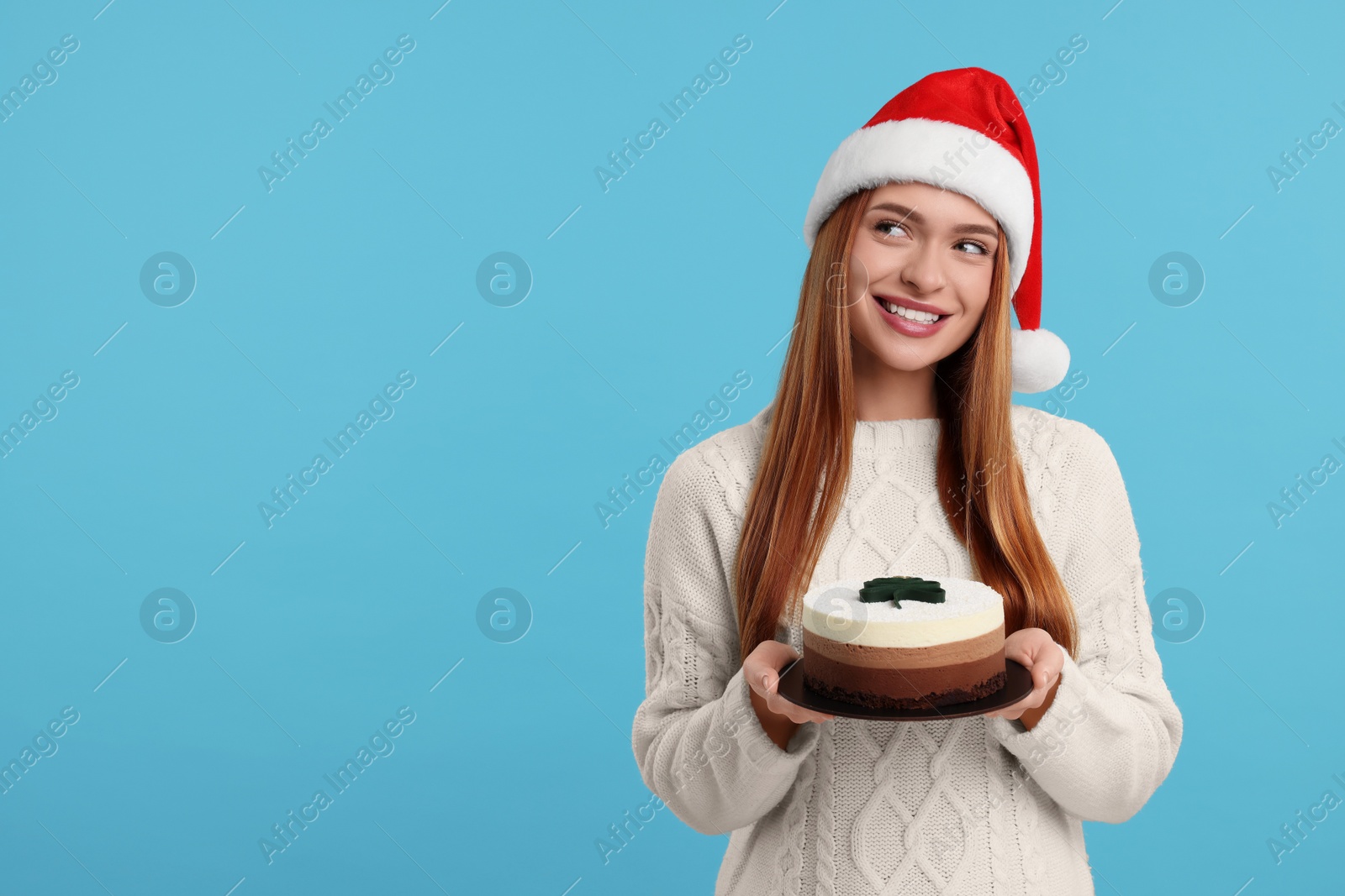 Photo of Young woman in Santa hat with tasty cake on light blue background, space for text