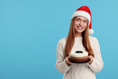 Young woman in Santa hat with tasty cake on light blue background, space for text