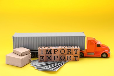 Photo of Words Import and Export made of wooden cubes near toy truck, banknotes and boxes on yellow background