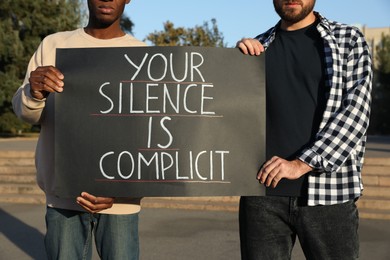 Photo of Men holding sign with phrase Your Silence Is Complicit outdoors, closeup. Racism concept