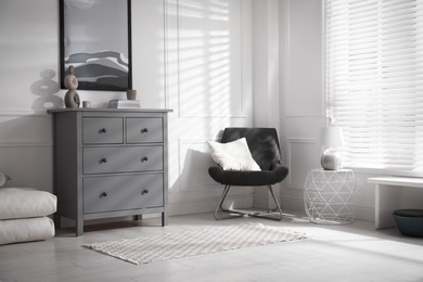 Stylish room interior with grey chest of drawers and comfortable chair