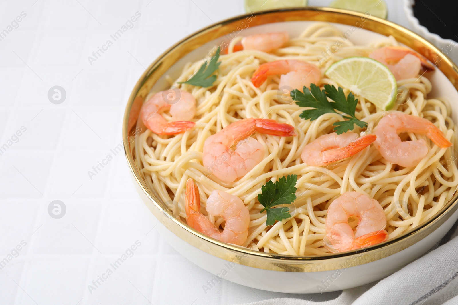 Photo of Tasty spaghetti with shrimps, lime and parsley in bowl on white tiled table, closeup. Space for text