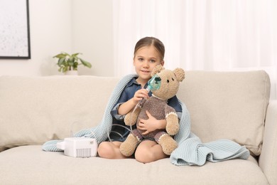 Photo of Little girl with toy bear and nebulizer for inhalation on sofa at home