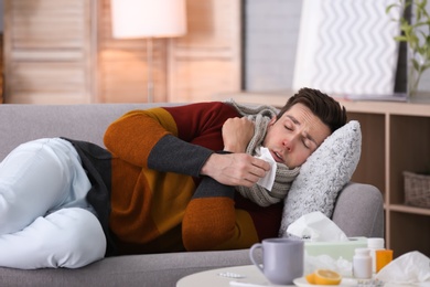 Sick young man with tissue suffering from cold on sofa at home