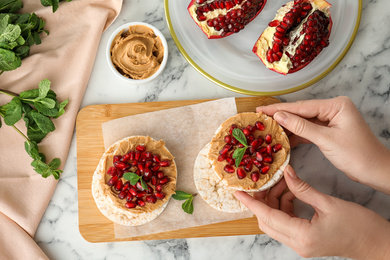 Photo of Woman holding puffed rice cake with peanut butter and pomegranate seeds at white marble table, top view