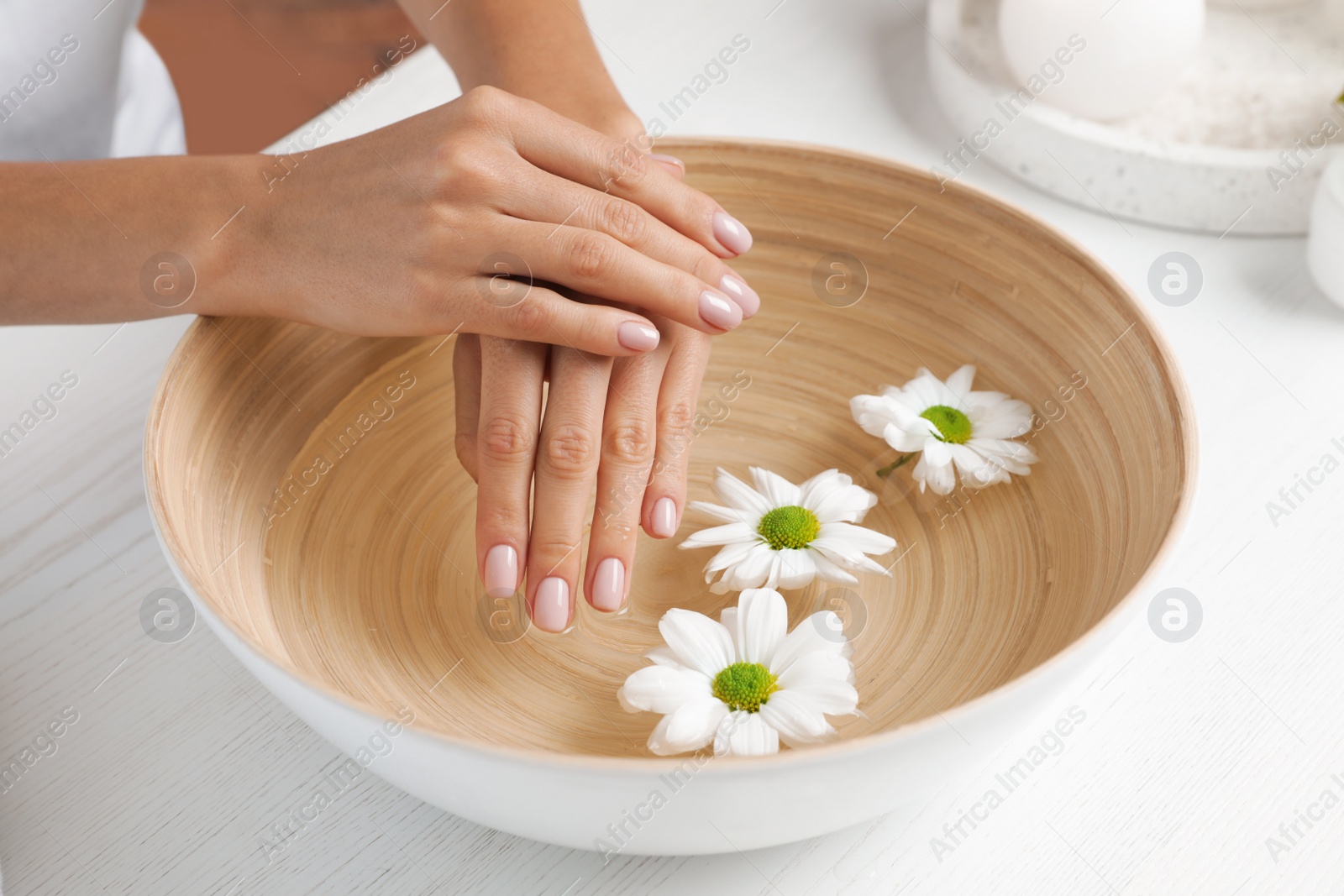 Photo of Woman soaking her hands in bowl with water and flowers on table, closeup. Spa treatment