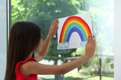 Photo of Little girl with picture of rainbow near window indoors.  Stay at home concept