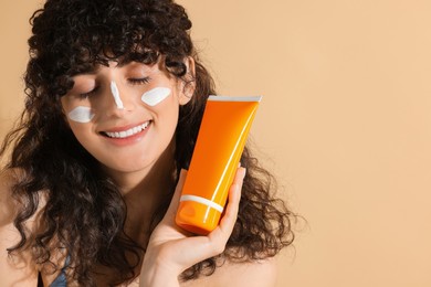 Photo of Beautiful young woman with sun protection cream on her face holding tube of sunscreen against beige background, space for text
