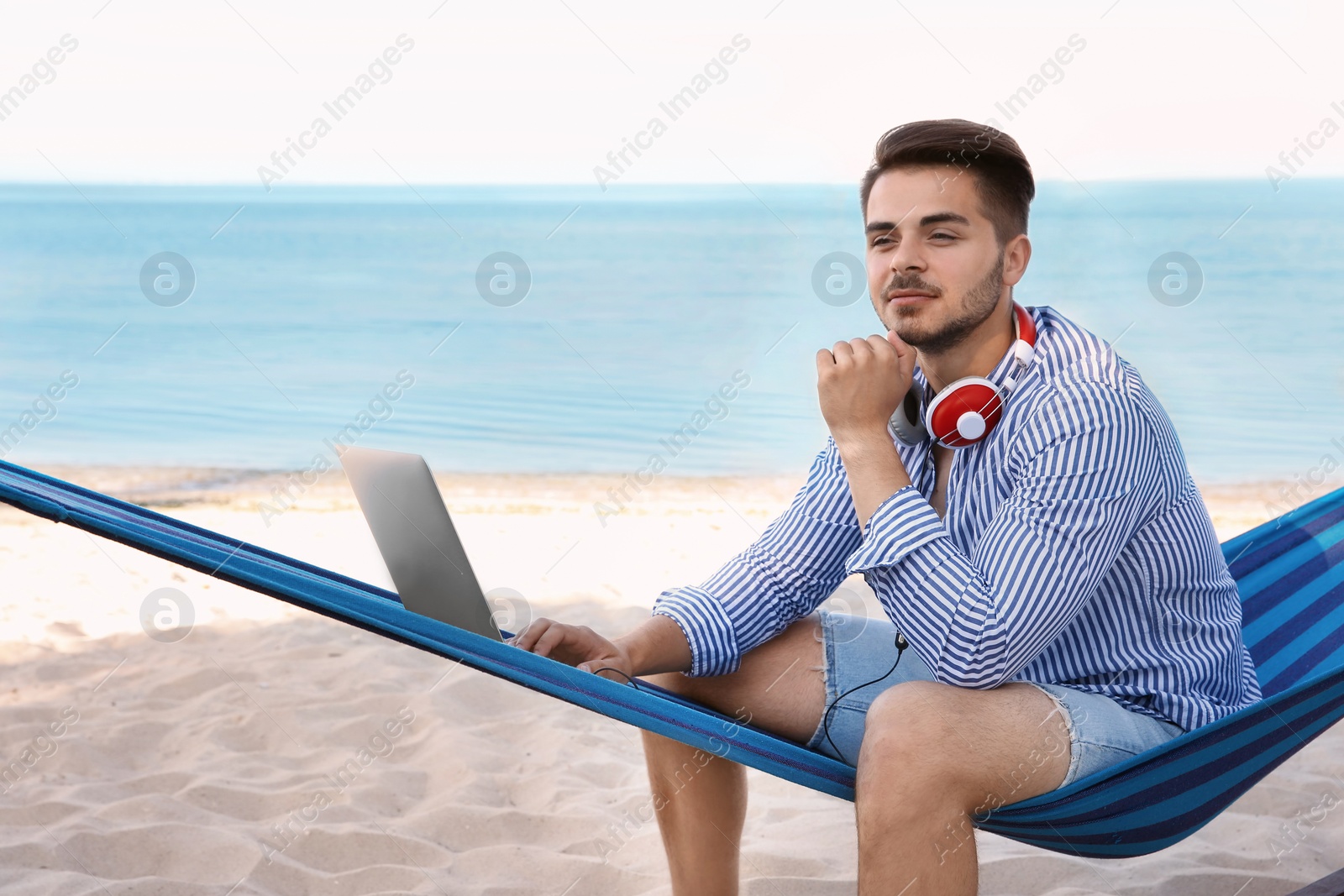 Photo of Young man with laptop and headphones sitting in hammock at seaside