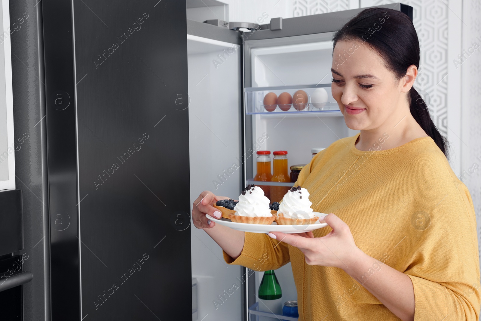 Photo of Happy overweight woman with cakes near fridge in kitchen