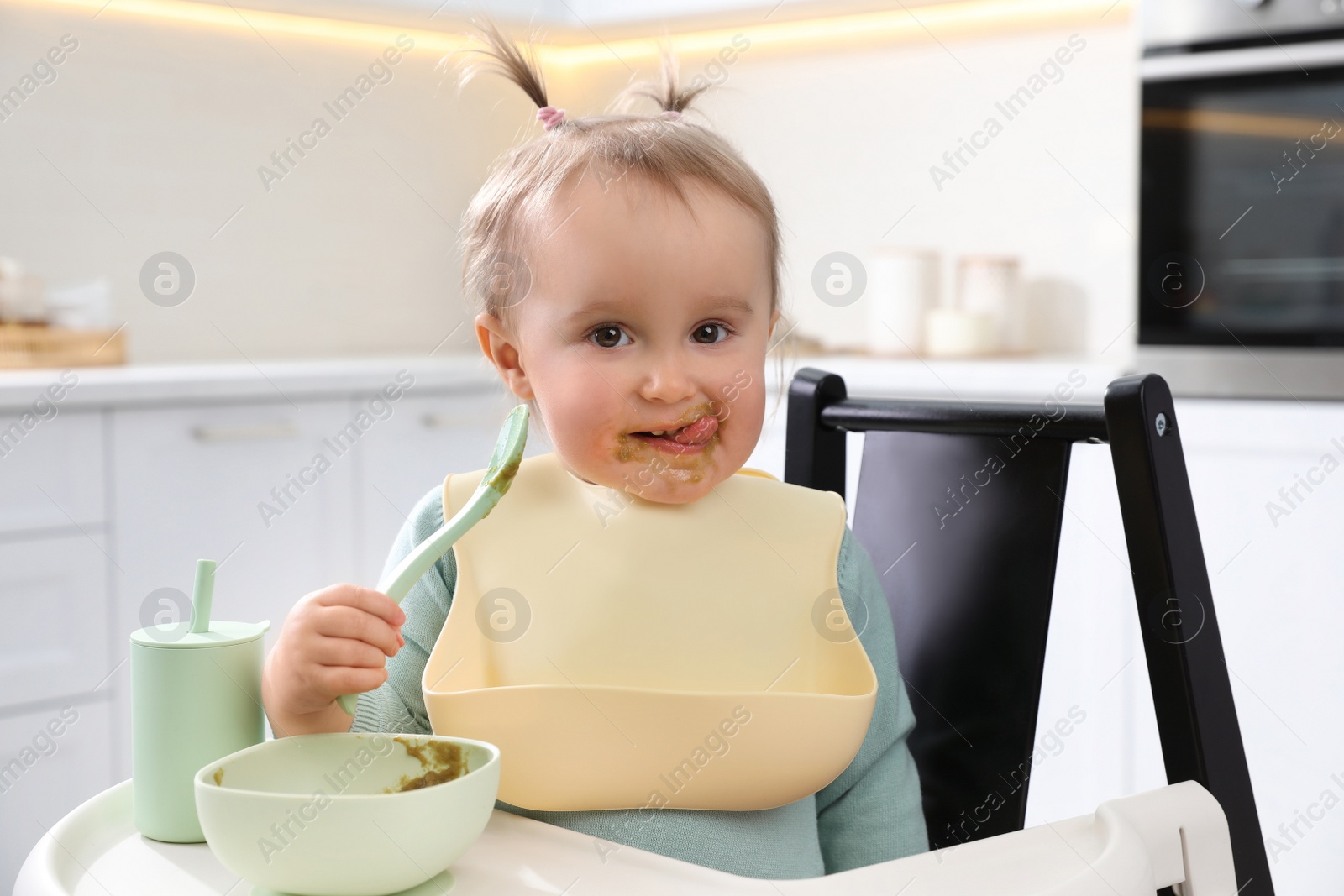 Photo of Cute little baby eating food in high chair at kitchen