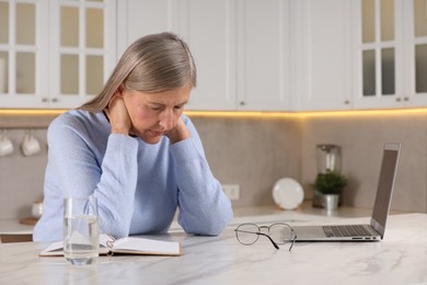 Menopause. Woman suffering from pain in neck at table in kitchen, space for text
