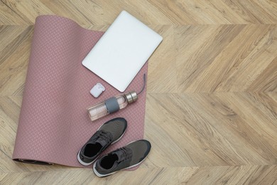 Photo of Exercise mat, laptop, bottle of water, wireless earphones and shoes on wooden floor, flat lay. Space for text