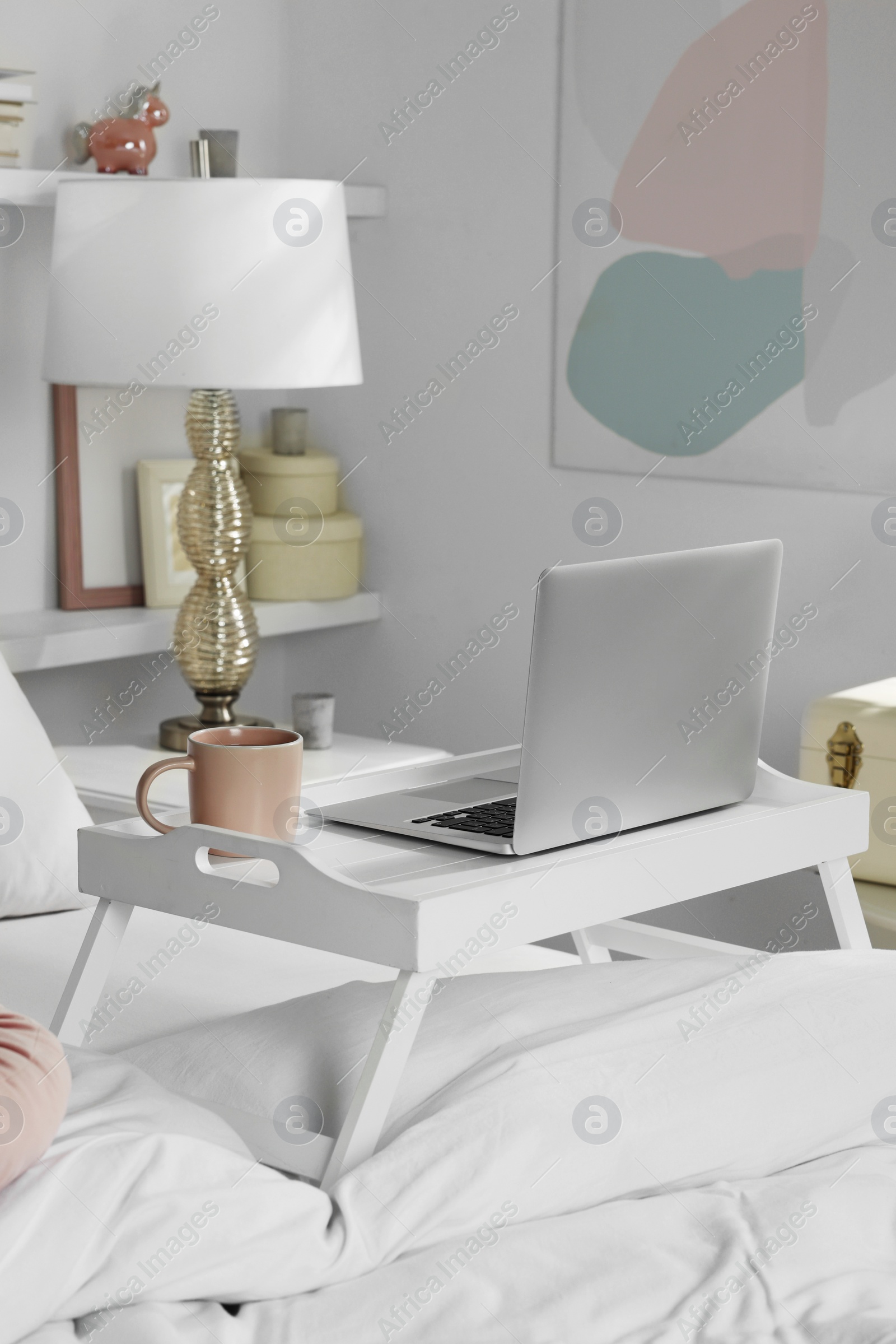 Photo of White tray table with laptop and cup of drink on bed indoors