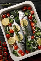 Photo of Raw fish with lemon in baking dish and vegetables on black wooden table, flat lay