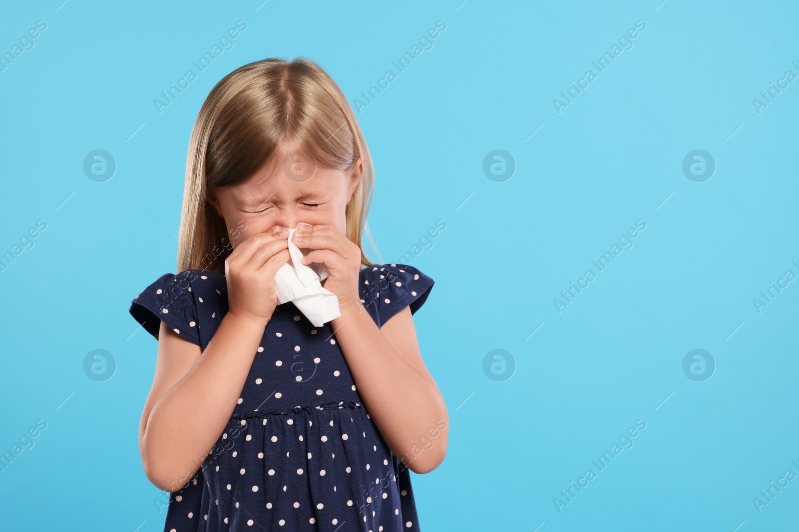 Photo of Suffering from allergy. Little girl with tissue sneezing on light blue background, space for text