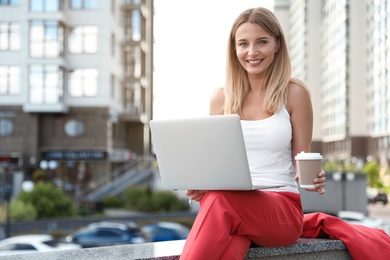Beautiful businesswoman with laptop and cup of coffee in city. Space for text