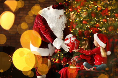 Photo of Santa Claus giving present to little girl near Christmas tree indoors