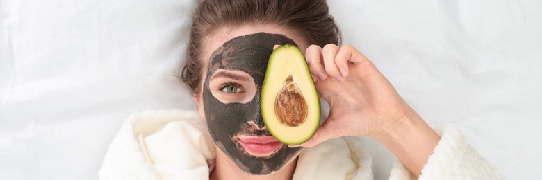 Image of Young woman with facial mask and avocado lying on bed, top view. Banner design