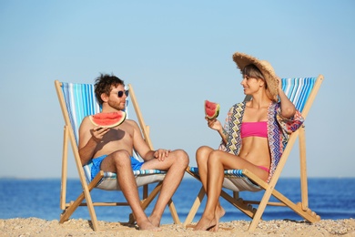 Young couple with watermelon slices in beach chairs at seacoast