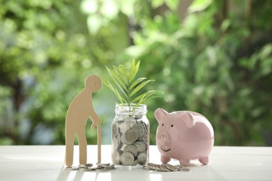 Pension savings. Figure of senior man, piggy bank, jar with coins and twig on white table against blurred green background