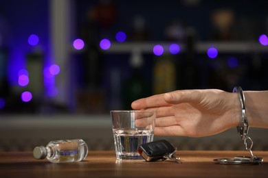 Man in handcuffs taking glass of alcohol near car key at wooden table indoors, closeup with space for text. Dangerous drinking and driving