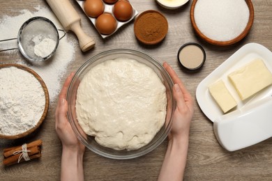 Woman holding bowl with fresh yeast dough and ingredients for cake on wooden table, top view