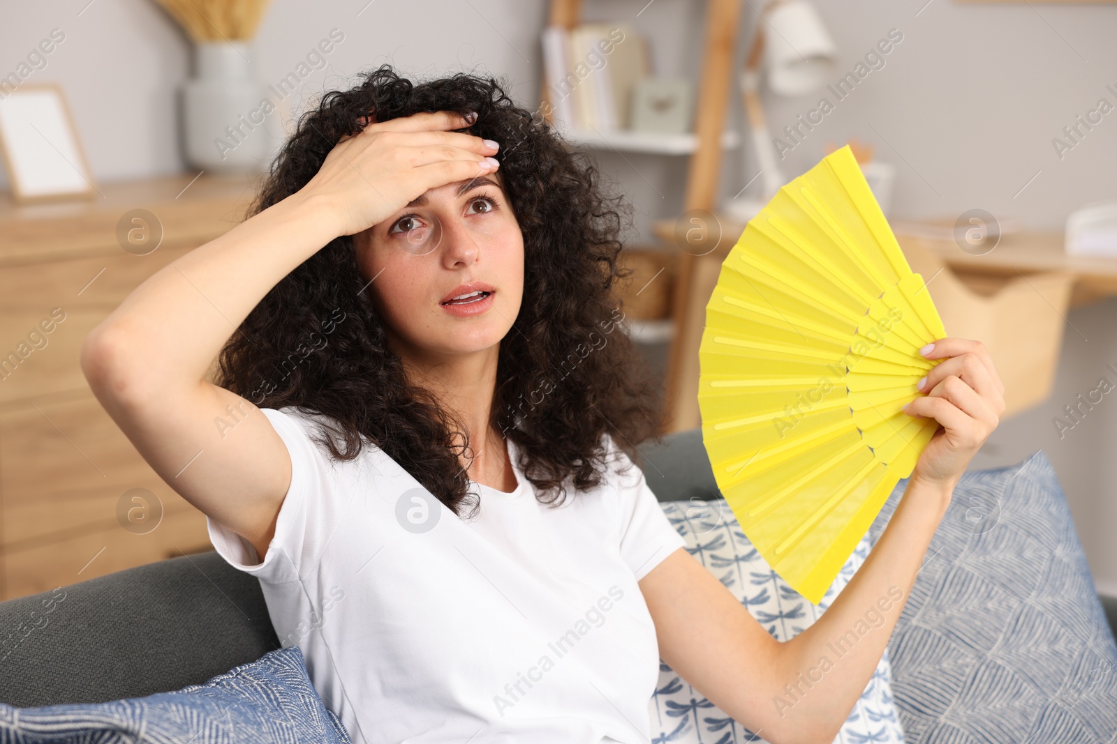 Photo of Young woman waving yellow hand fan to cool herself on sofa at home