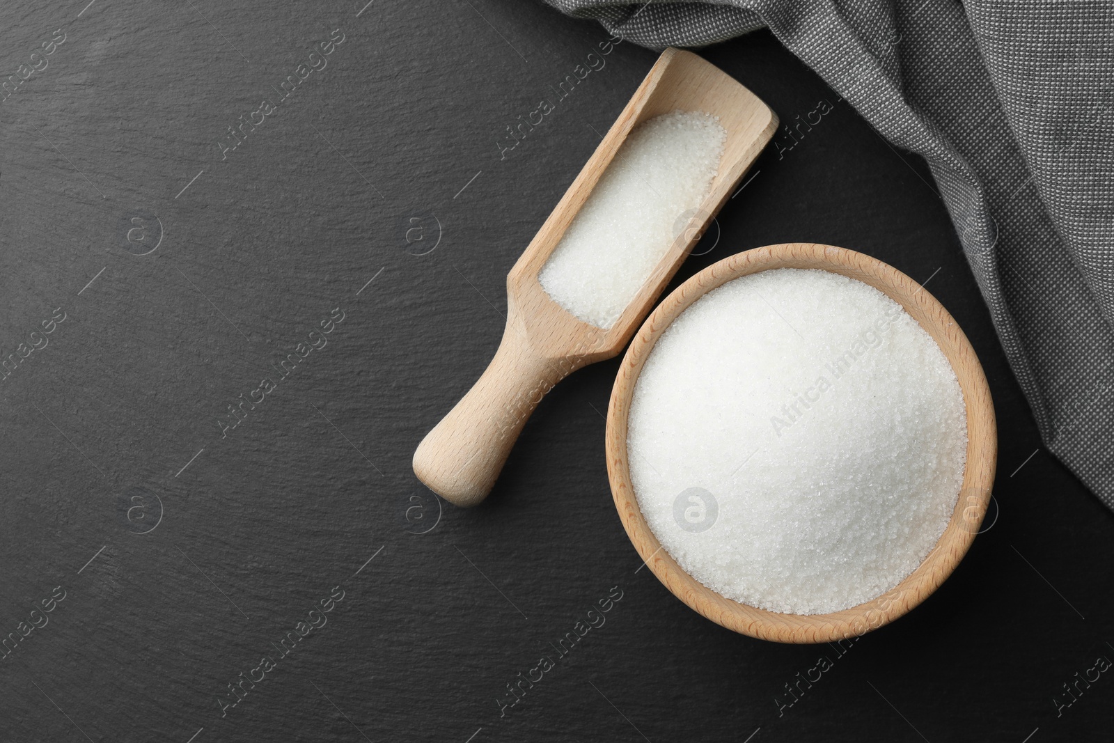 Photo of Granulated sugar in bowl and scoop on black table, flat lay. Space for text