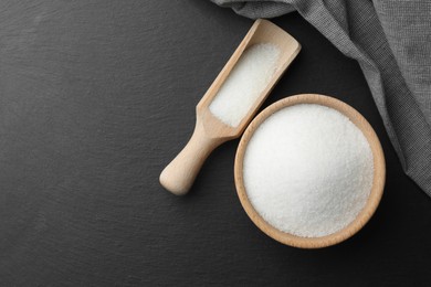 Granulated sugar in bowl and scoop on black table, flat lay. Space for text