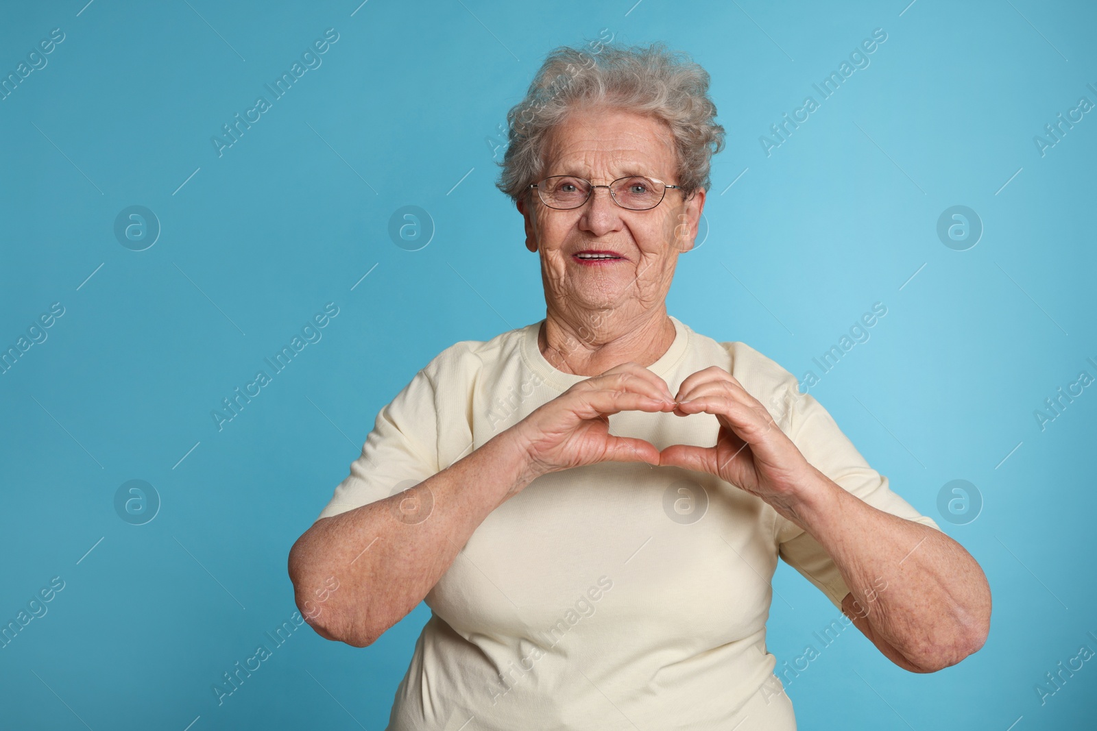 Photo of Elderly woman making heart with her hands on light blue background, space for text