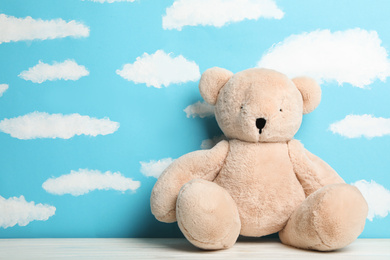 Photo of Teddy bear on white wooden table near wall with painted blue sky, space for text. Baby room interior
