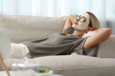 Photo of Young woman with face mask and cucumber slices resting on sofa at home. Spa treatments
