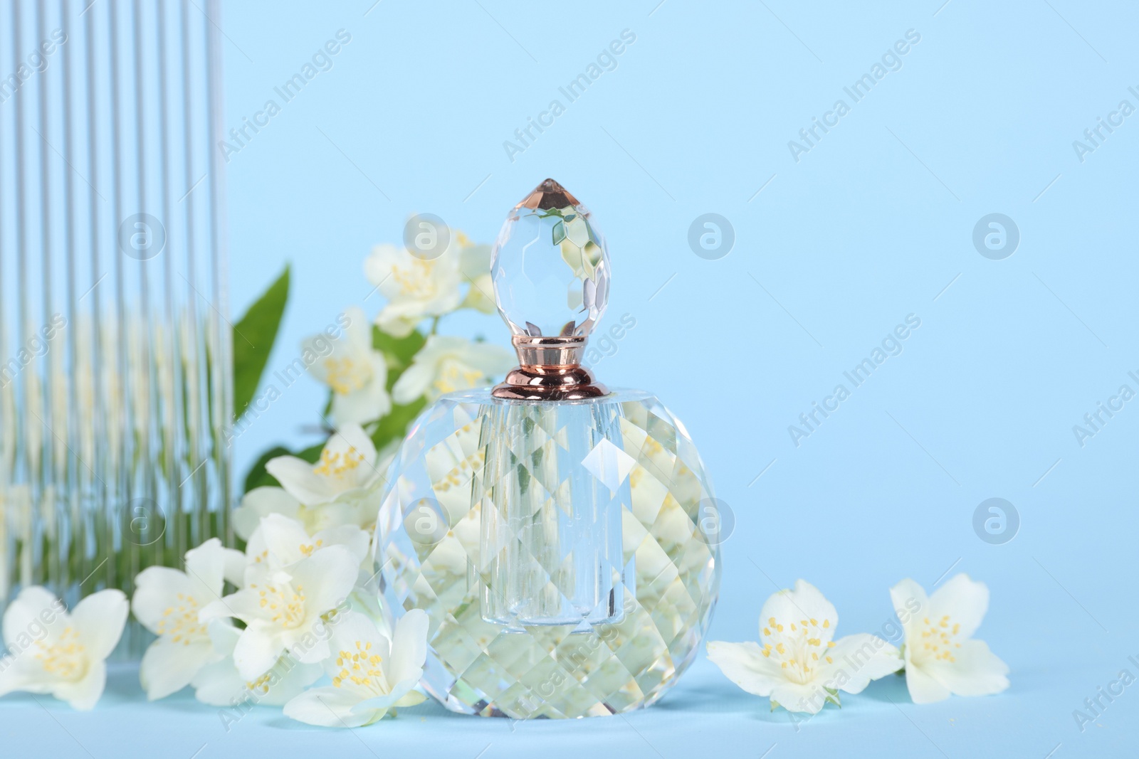 Photo of Aromatic perfume in bottle and beautiful jasmine flowers on light blue background, closeup. Space for text