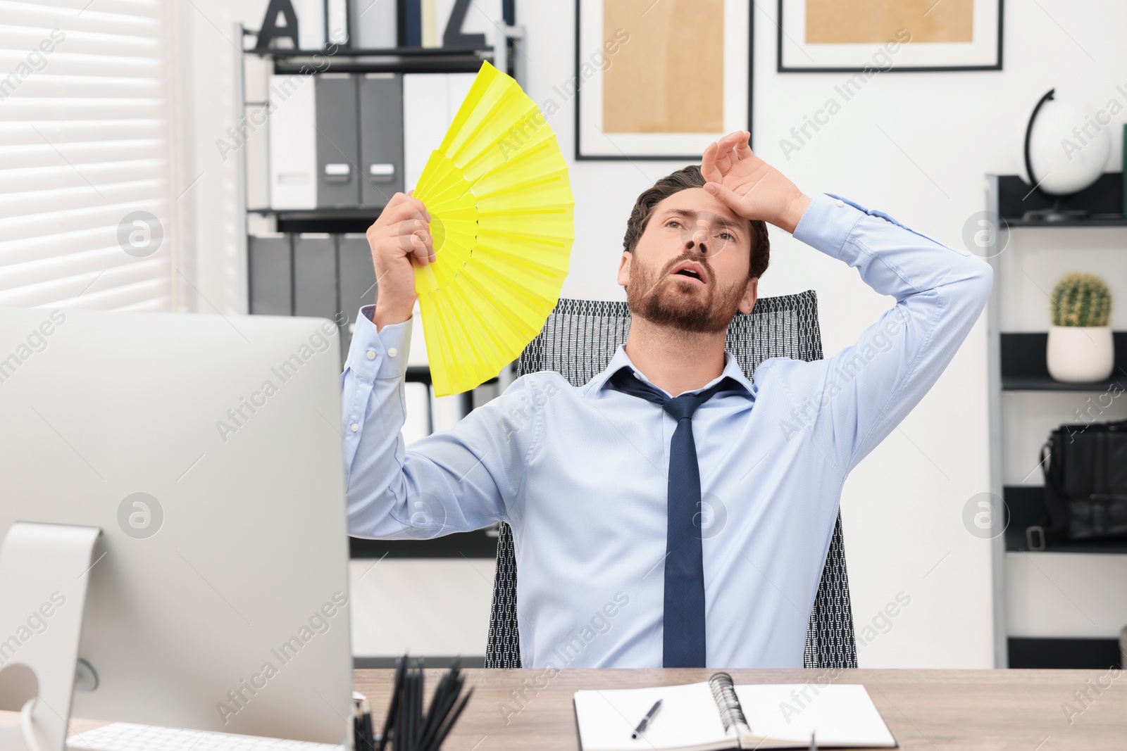 Photo of Bearded businessman waving yellow hand fan to cool himself at table in office