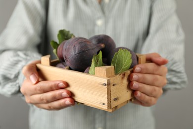 Woman holding wooden crate with tasty raw figs on light grey background, closeup