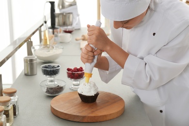 Young female pastry chef decorating cupcake with cream at table in kitchen