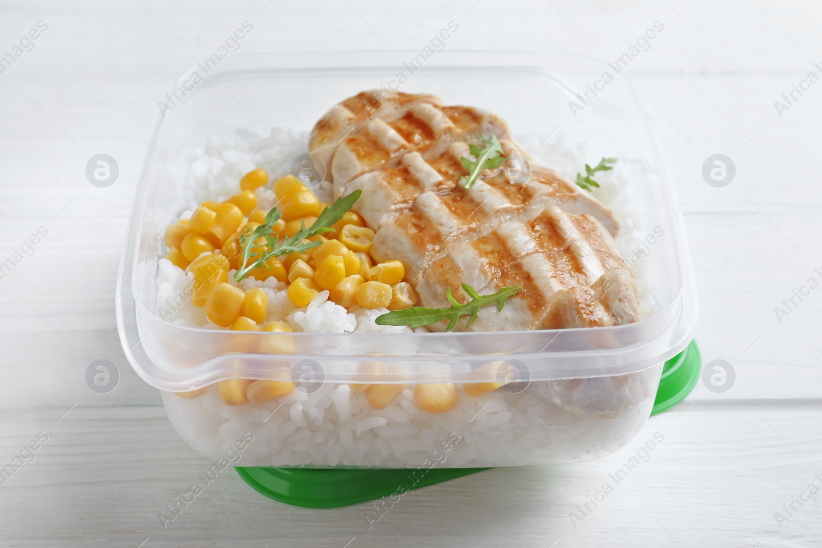 Photo of Tasty rice with grilled meat and corn in plastic container on white wooden table