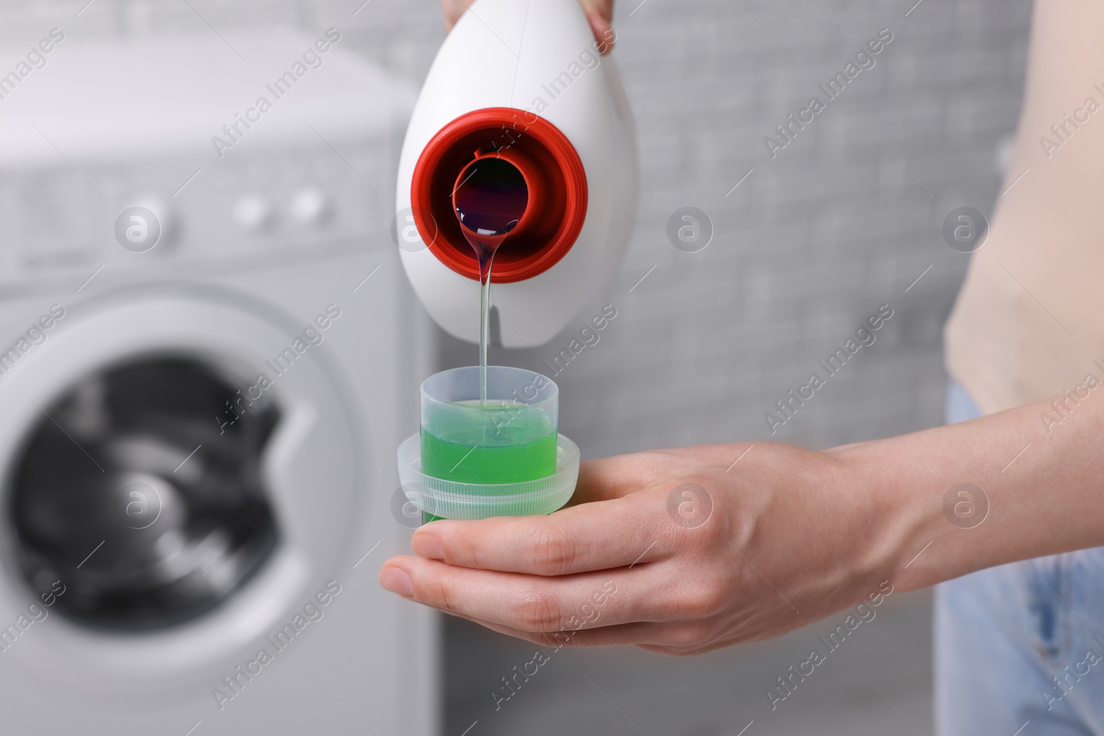 Photo of Woman pouring fabric softener from bottle into cap for washing clothes indoors, closeup