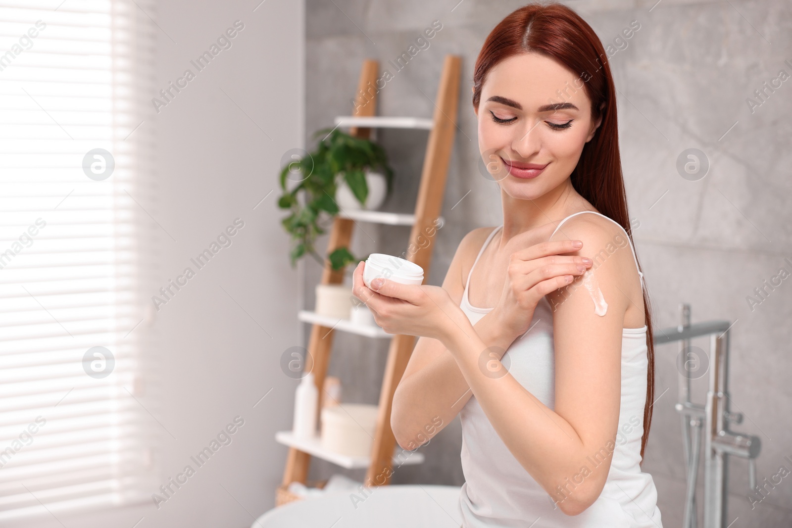 Photo of Beautiful young woman applying body cream onto shoulder in bathroom, space for text