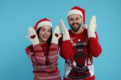 Photo of Young couple in Christmas sweaters, Santa hats and knitted mittens on light blue background