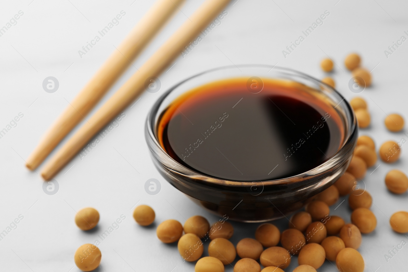 Photo of Soy sauce in bowl and soybeans on white table, closeup