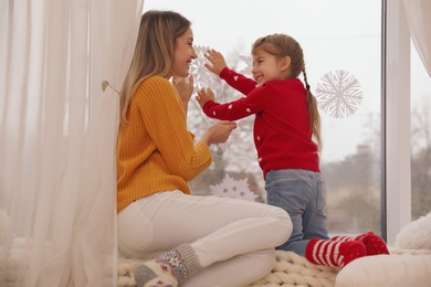 Happy mother and daughter decorating window with paper snowflakes indoors