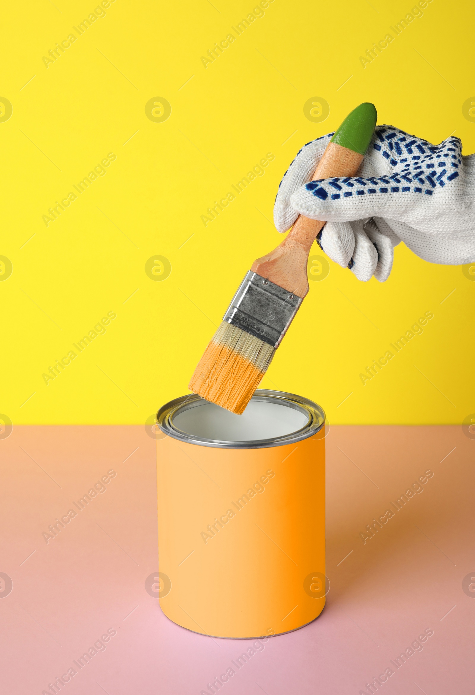 Photo of Person dipping brush into can of orange paint on pink table against yellow background, closeup. Mockup for design