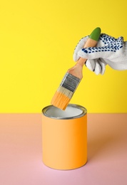 Photo of Person dipping brush into can of orange paint on pink table against yellow background, closeup. Mockup for design