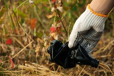 Photo of Woman in gloves picking crumpled bottle from grass outdoors, closeup. Space for text