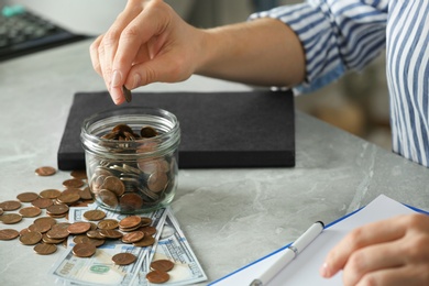 Photo of Woman putting coin into jar at grey marble table, closeup. Money savings