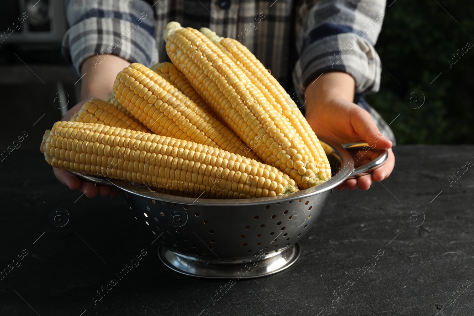 Photo of Woman with colander of corn cobs at black table, closeup