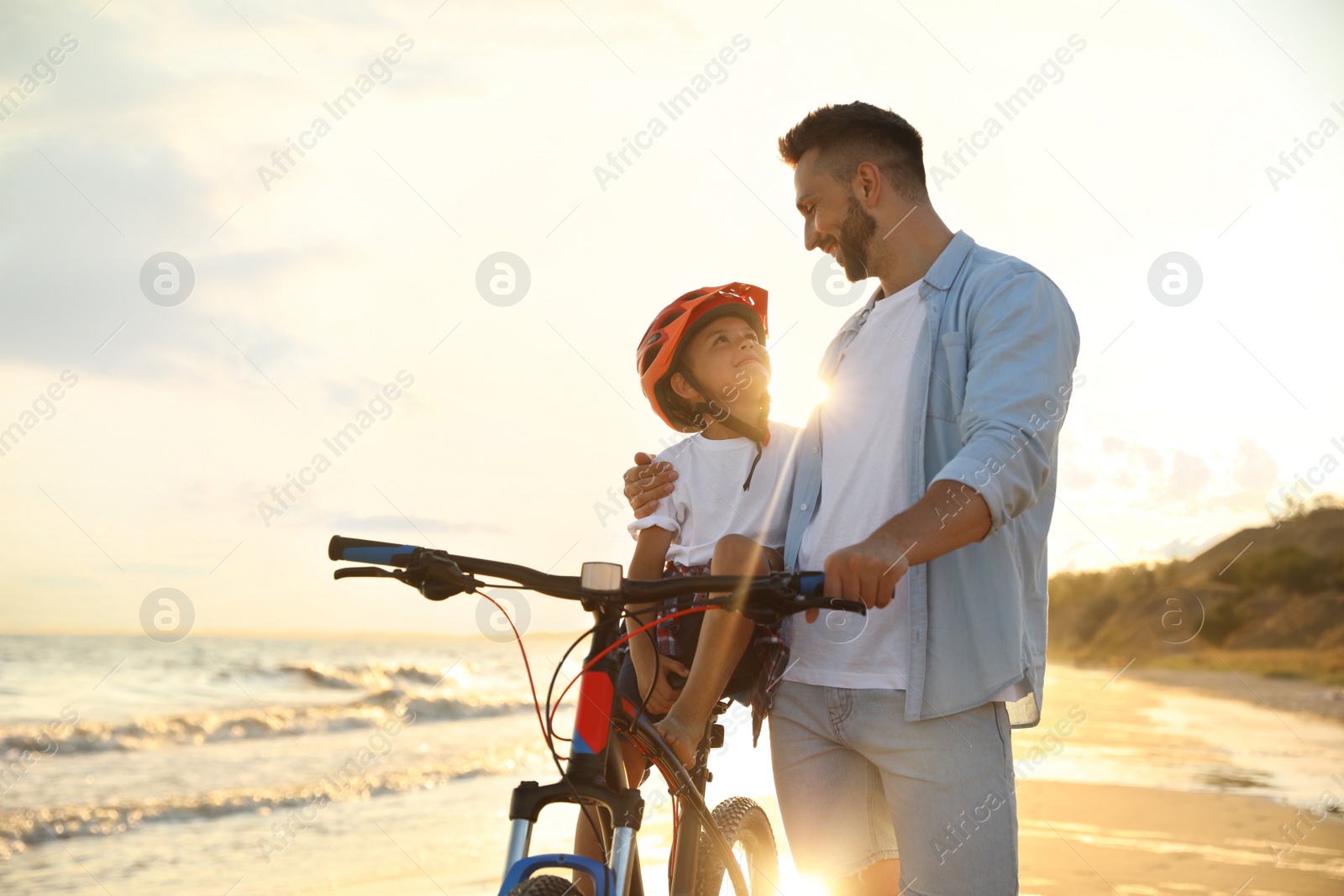 Photo of Happy father teaching son to ride bicycle on sandy beach near sea at sunset